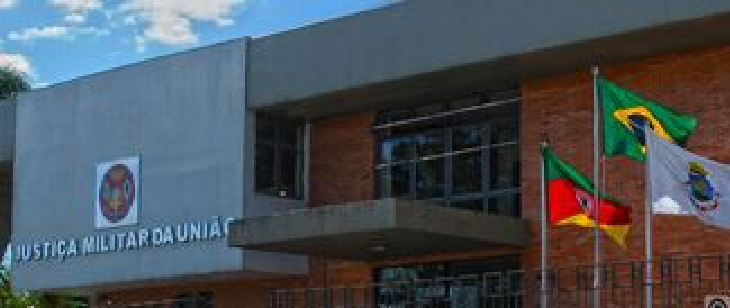 Fachada da 3ª CJM – unidade de Bagé. Prédio com fachada de tijolos vermelhos e estrutura de concreto. Na parte frontal, três bandeiras estão hasteadas: a do Brasil, a do Rio Grande do Sul e a institucional. O céu azul e as árvores completam o cenário externo.